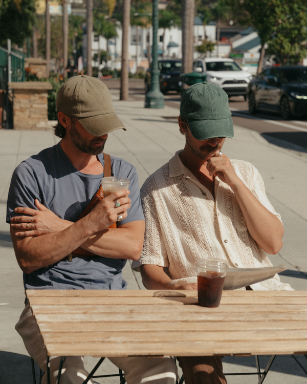 Two models wearing blank khaki and dark green Classic Caps at an outdoor cafe
